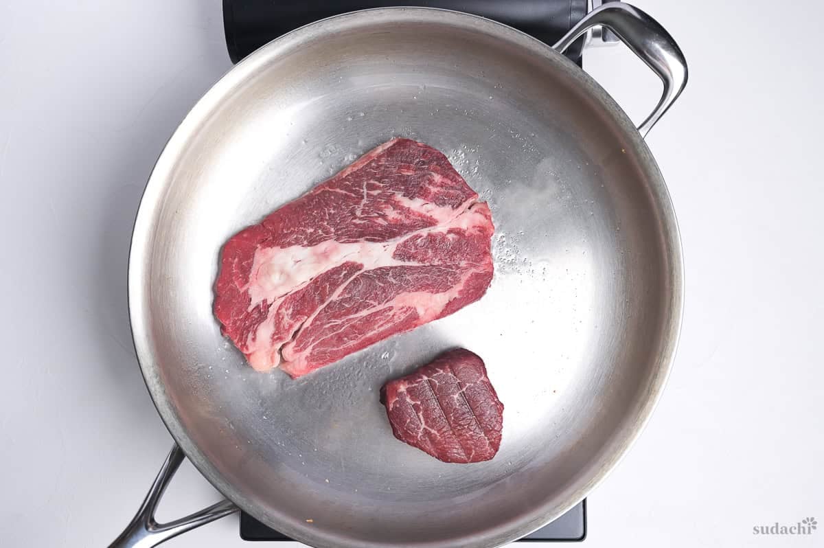 Beef steaks frying in a large stainless steel pan on the stove top.
