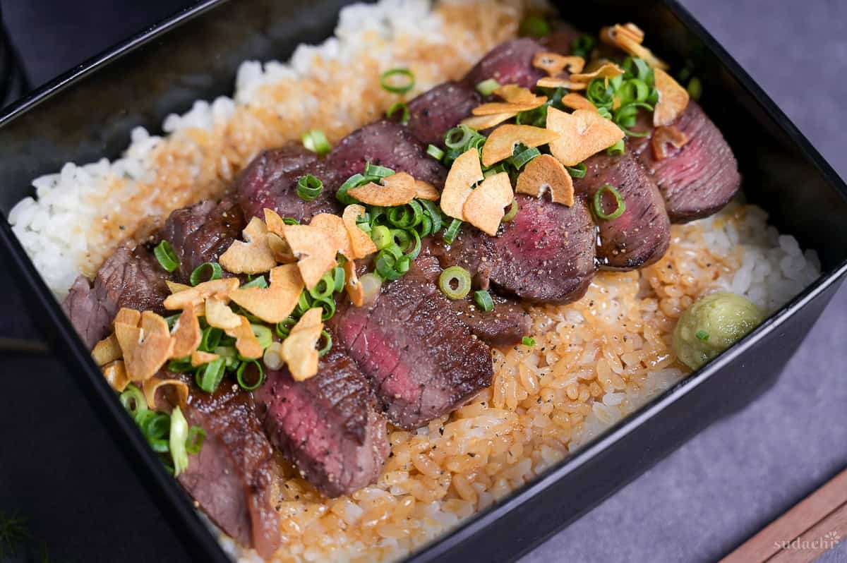 Steak Jyu (Japanese steak rice in a box) topped with chopped green onion, crispy garlic and ground black pepper with a blob of wasabi served in a black jubako.