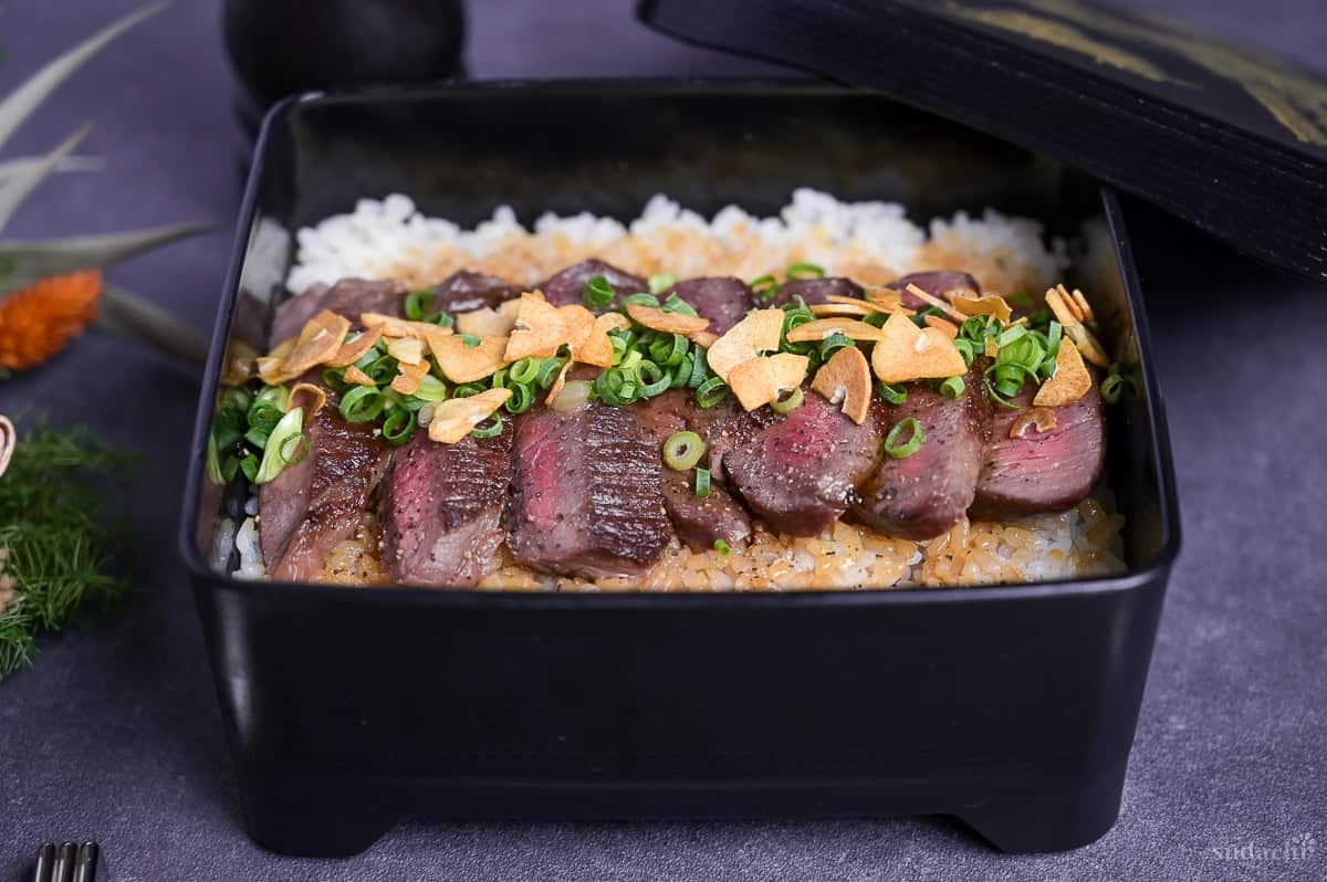 Steak Jyu (Japanese steak rice in a box) topped with chopped green onion, crispy garlic and ground black pepper with a blob of wasabi served in a black jubako.