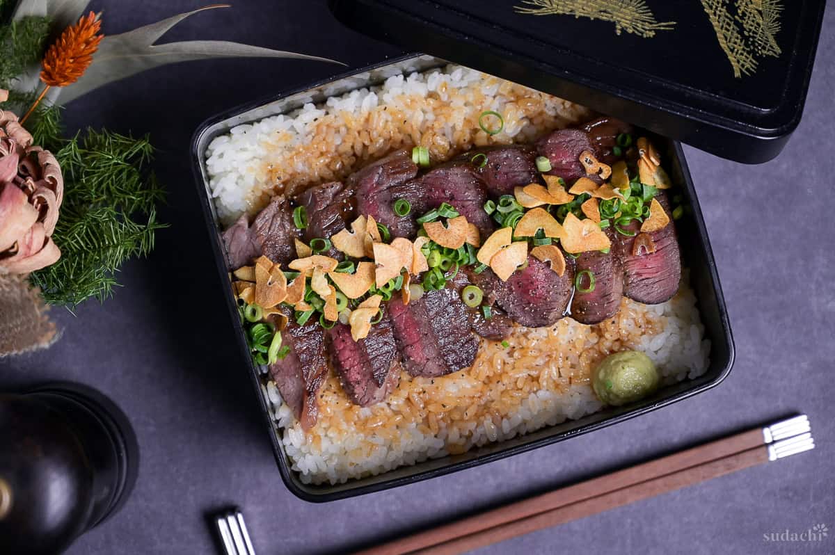 Steak Jyu (Japanese steak rice in a box) topped with chopped green onion, crispy garlic and ground black pepper with a blob of wasabi served in a black jubako.