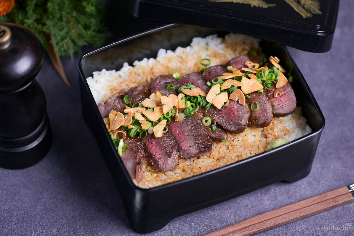Steak Jyu (Japanese steak rice in a box) topped with chopped green onion, crispy garlic and ground black pepper with a blob of wasabi served in a black jubako.