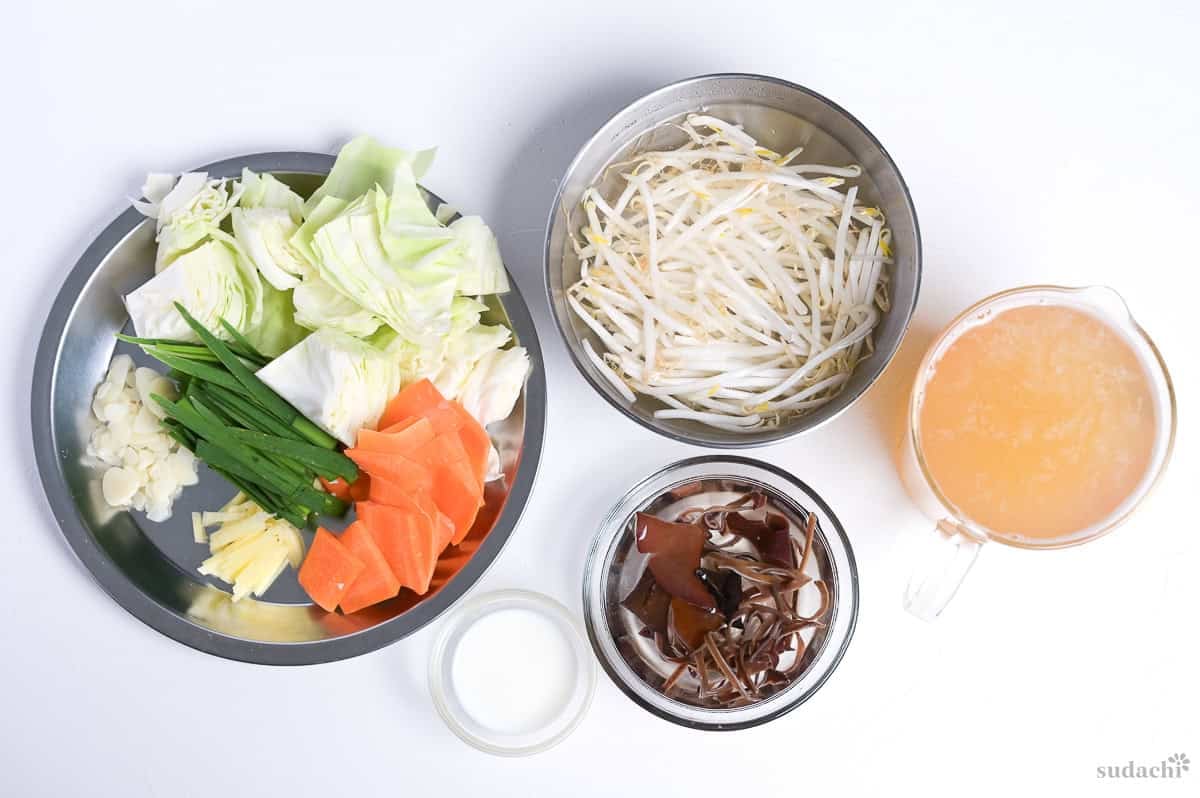 Ingredients prepared for tanmen. Left plate: roughly cut cabbage, thinly sliced garlic cloves, ginger root and carrot, sliced garlic chives. Middle bowls: bean sprouts in water, milk in a small bowl, wood ear mushrooms rehydrating in water. Glass jug on the right: chicken bouillon powder and condiments mixed with hot water.