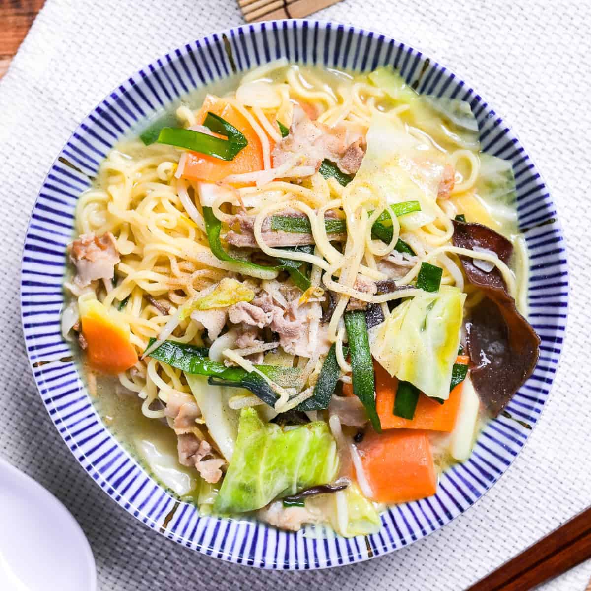 Japanese tanmen noodles in a white bowl with blue striped edge on a woven placemat.