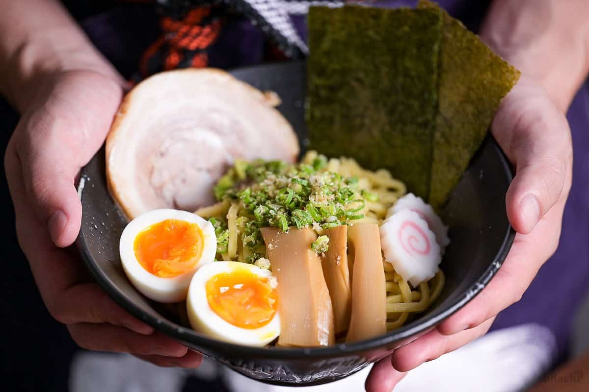 Yuto Omura holding up a black bowl of abura soba topped with soft boiled eggs, menma, narutomaki, nori, pork chashu and chopped green onions.