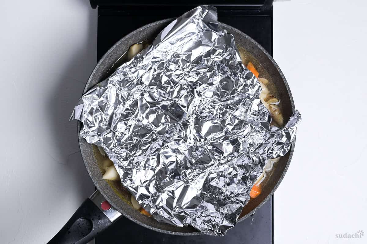chikuzenni simmering in a wide pan on the stove top with drop lid made with foil