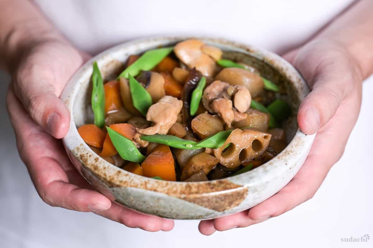 Yuto Omura holding up a bowl of chikuzenni made with chicken thigh pieces, carrot, lotus root, burdock root, and shiitake mushroom simmered in a dashi based broth.