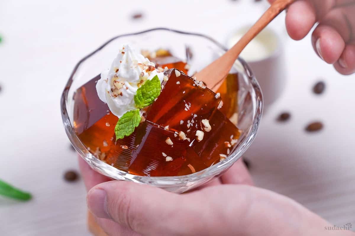 Cubes of Japanese coffee jelly topped with whipped cream, chopped nuts and a mint leaf served in a glass bowl with a wooden dessert fork taking a cube