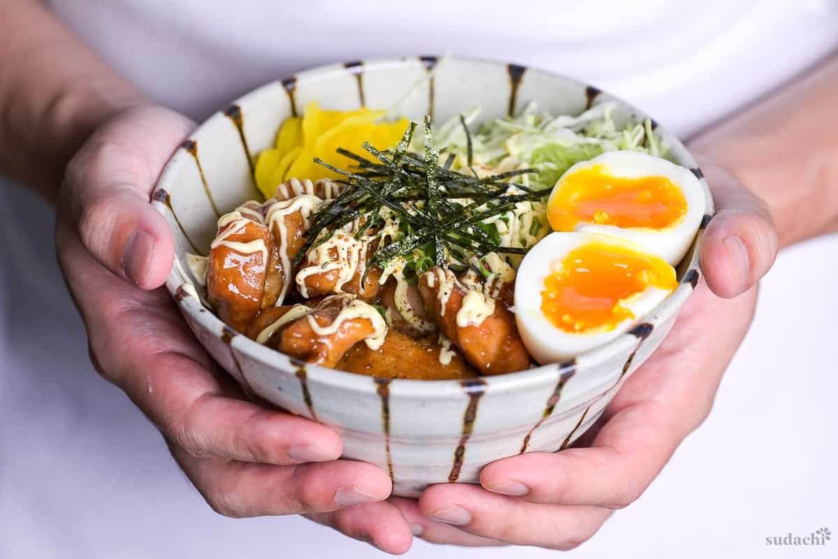 Yuto Omura holding garlic teriyaki chicken donburi with shredded cabbage, soft boiled eggs, takuan pickles and nori in an off-white and brown striped bowl