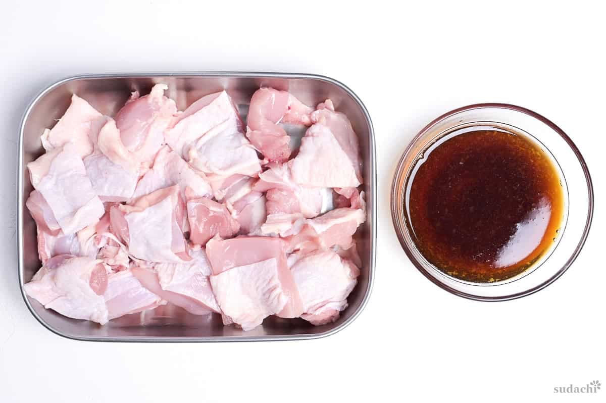 raw chicken thigh cut into large pieces and placed in a steel container next to teriyaki sauce in a small glass bowl