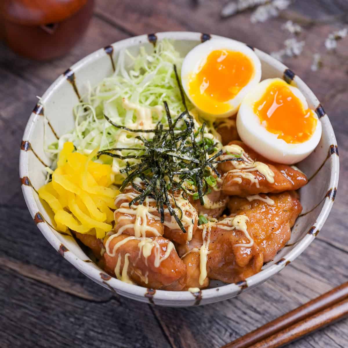 Garlic teriyaki chicken donburi with shredded cabbage, soft boiled eggs, takuan pickles and nori served in a striped bowl