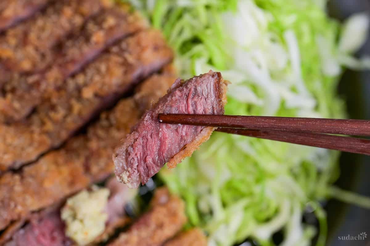 A close up of a piece of gyukatsu held up with wooden chopsticks