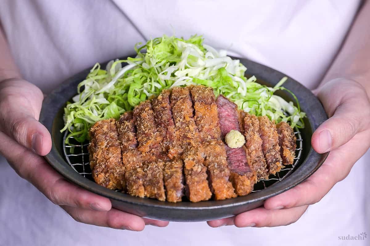 Yuto Omura holding up Gyukatsu (Japanese beef cutlet) cooked rare and served on a wire rack with shredded cabbage