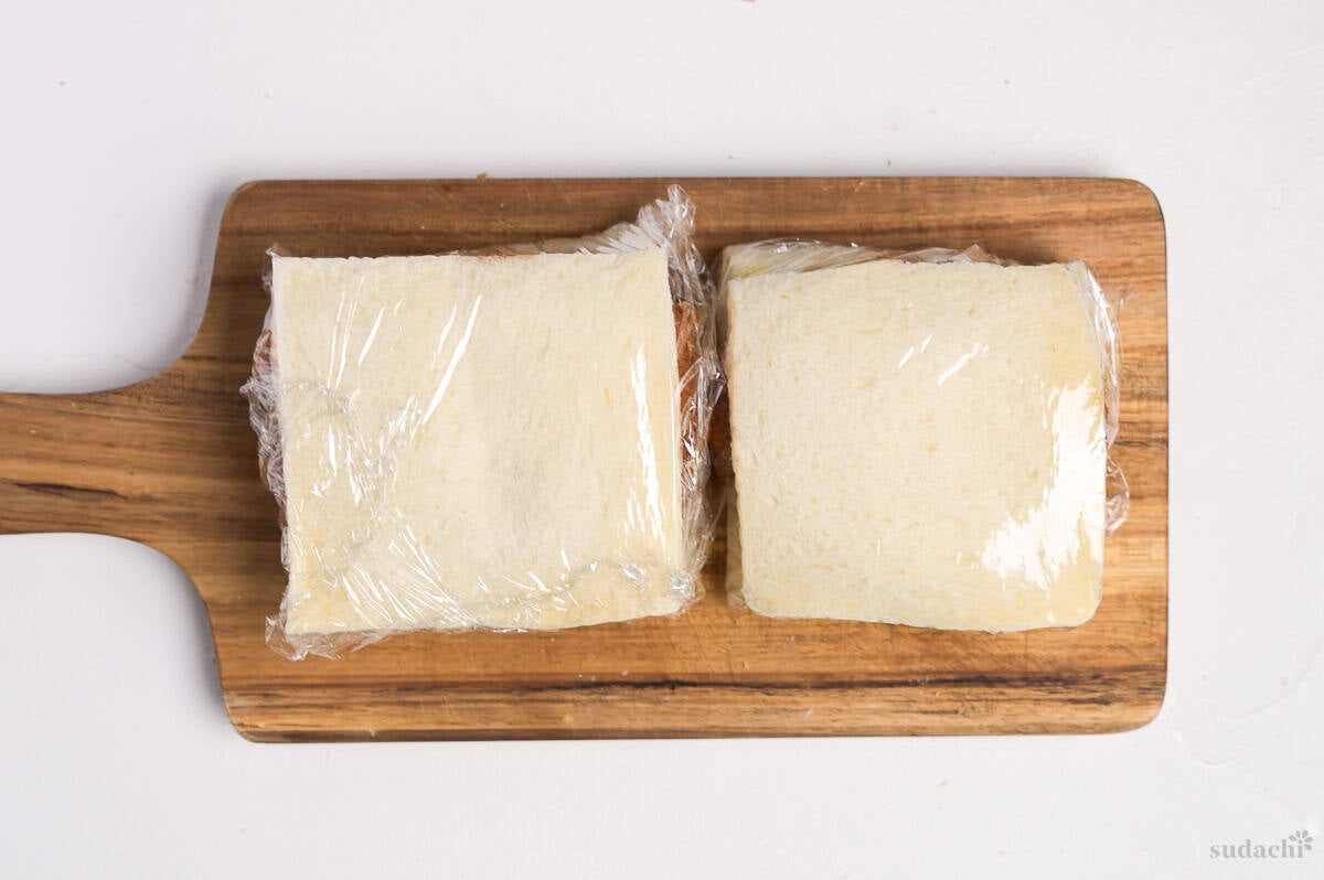 A katsu sando (Japanese pork cutlet sandwich) wrapped with plastic wrap on a wooden cutting board