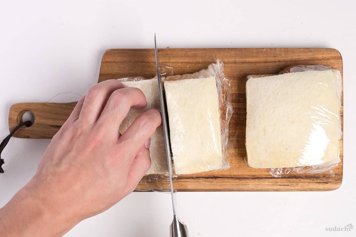 A katsu sando (Japanese pork cutlet sandwich) wrapped with plastic wrap and being cut in half on a wooden cutting board