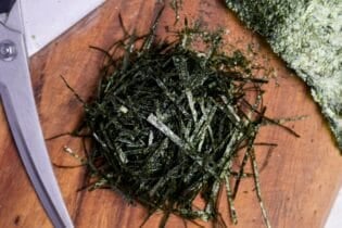 hand cut kizami nori on a wooden cutting board next to a pair of scissors and a few whole sheets of yaki nori