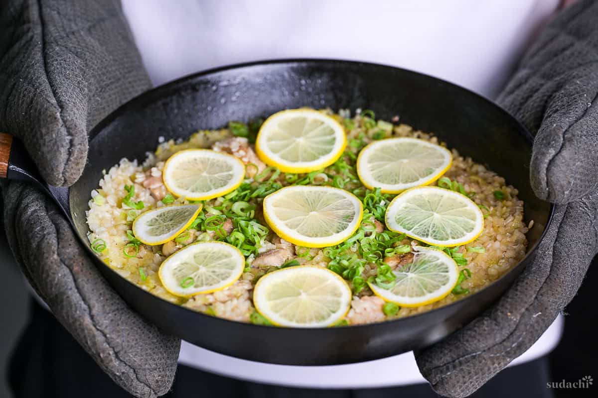 Yuto Omura holding up Japanese style negi shio paella with chicken and lemon in a black frying pan topped with lemon slices and finely chopped green onion
