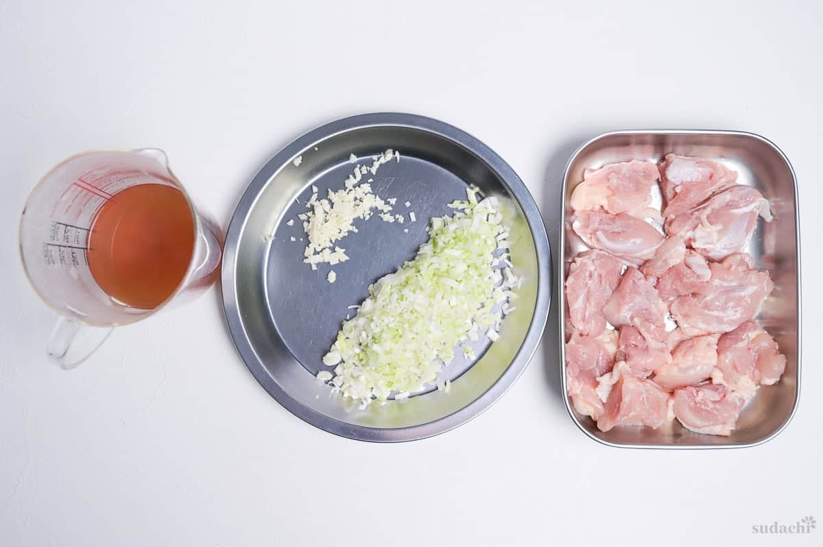 Stock for negi shio paella in a glass measuring jug, finely chopped garlic and Japanese leek on a silver prep plate, and chicken thigh cut into bitesize pieces in a steel container.