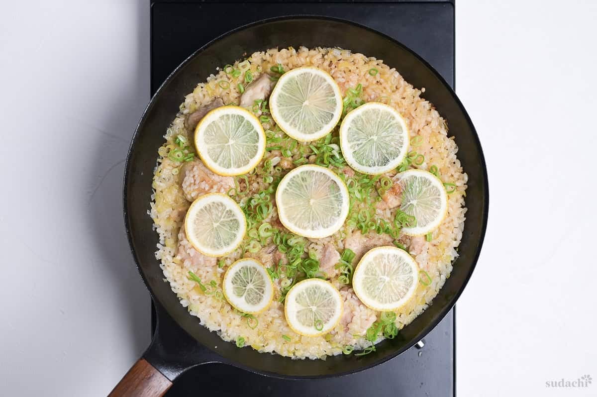 Japanese style negi shio paella with chicken and lemon in a black frying pan topped with lemon slices and finely chopped green onion on the stove top