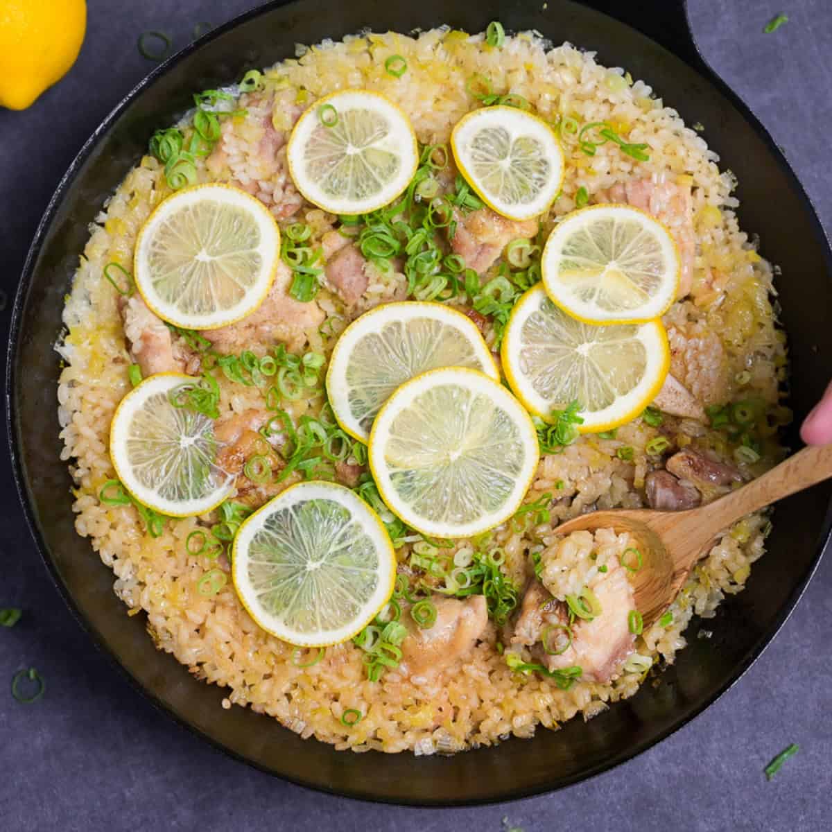 Japanese style negi shio paella with chicken and lemon in a black frying pan topped with lemon slices and finely chopped green onion with wooden spoon scooping (top down)