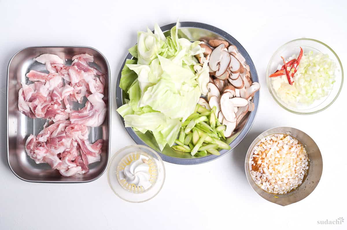 Ingredients prepped for making negishio spaghetti. From left to right: thinly sliced pork belly in a steel container, squeezed lemon juice, roughly cut cabbage, sliced shiitake and asparagus, finely diced aromatics, negishio sauce.
