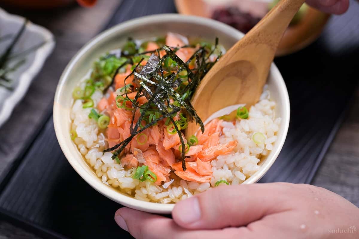 ochazuke in a bowl topped with salmon flakes, chopped green onions and kizami nori being scooped with a wooden spoon