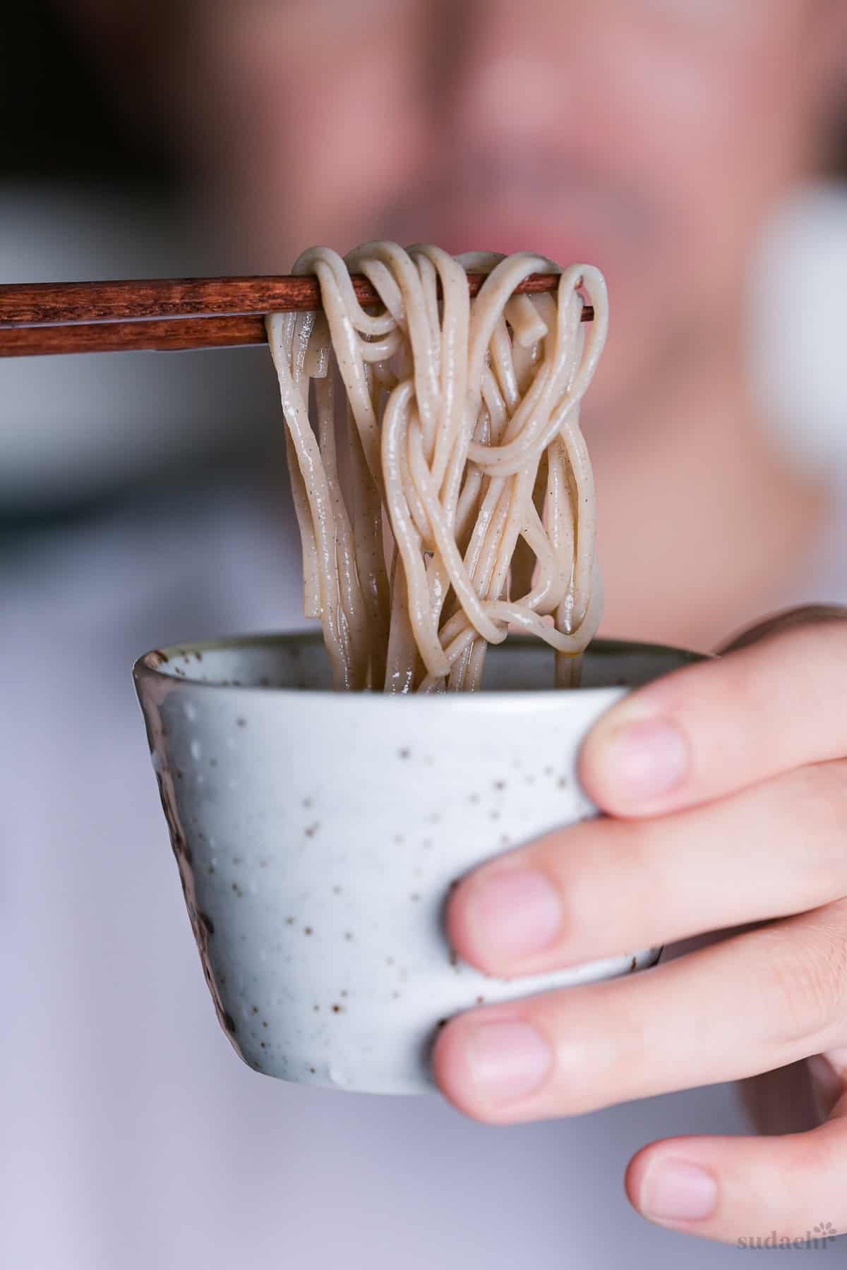 Yuto Omura holding up zaru soba, chilled homemade mentsuyu in a mottled dipping cup with chilled soba noodles held above with wooden chopsticks.