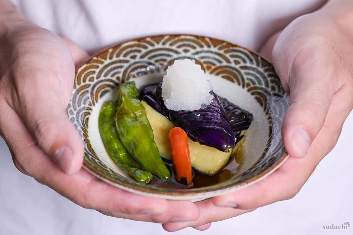 Yuto Omura holding nasu no agebitashi (Fried Japanese eggplant in dashi) with shishito peppers and chili topped with grated daikon radish in a dish with brown and grayish blue seigaiha pattern