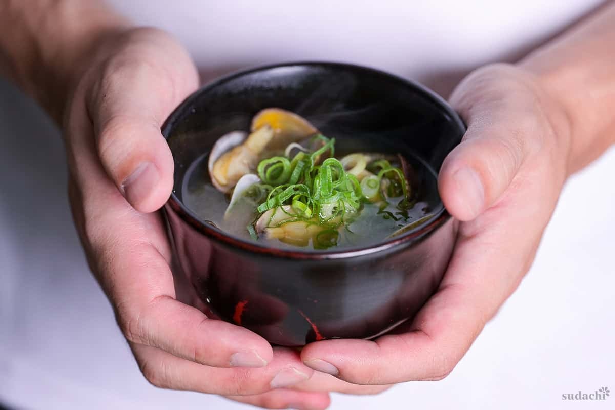 Yuto Omura holding homemade asari (clam) miso soup topped with green onions served in a small black Japanese soup bowl