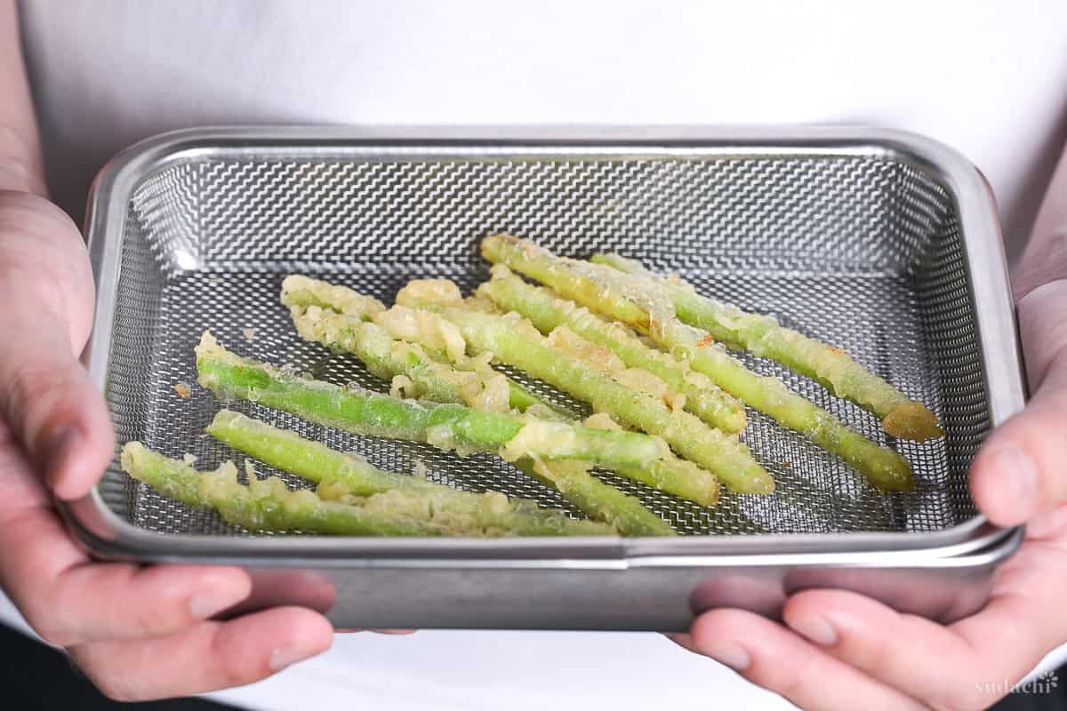 Yuto Omura holding asparagus tempura on a mesh wire rack inside a steel container.