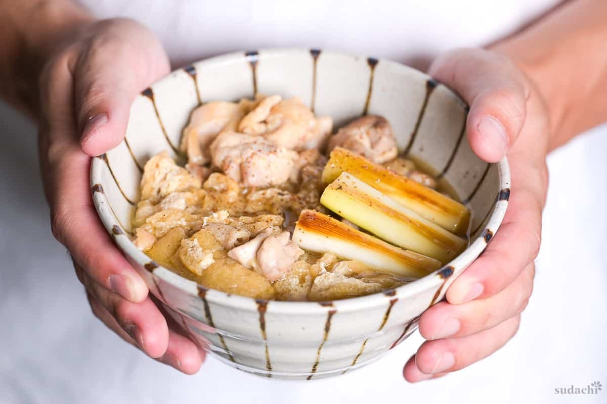 Yuto Omura holding up a bowl of chicken udon noodle soup (tori nanban udon) topped with grilled Japanese leek, pan fried chicken thigh and aburaage (fried tofu pouch) in a striped bowl