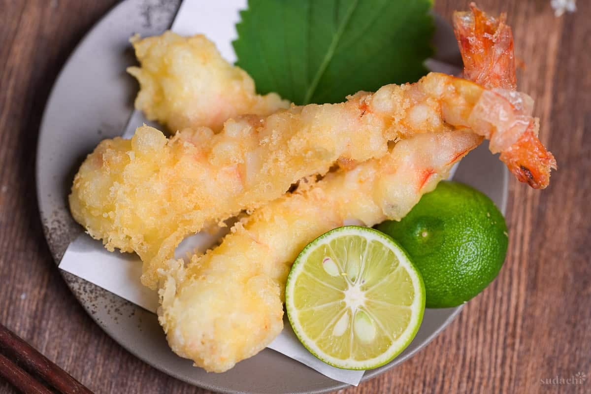 Three homemade ebiten (shrimp tempura) arranged on a gray plate next to a halved sudachi and shiso leaf top down