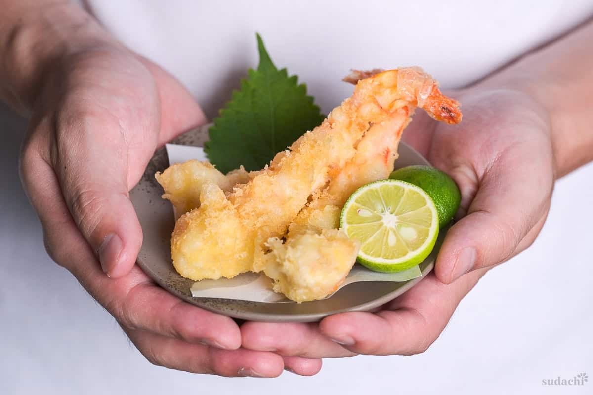 Yuto Omura holding up ebi tempura (shrimp) with sudachi and perilla leaf on a brownish gray plate