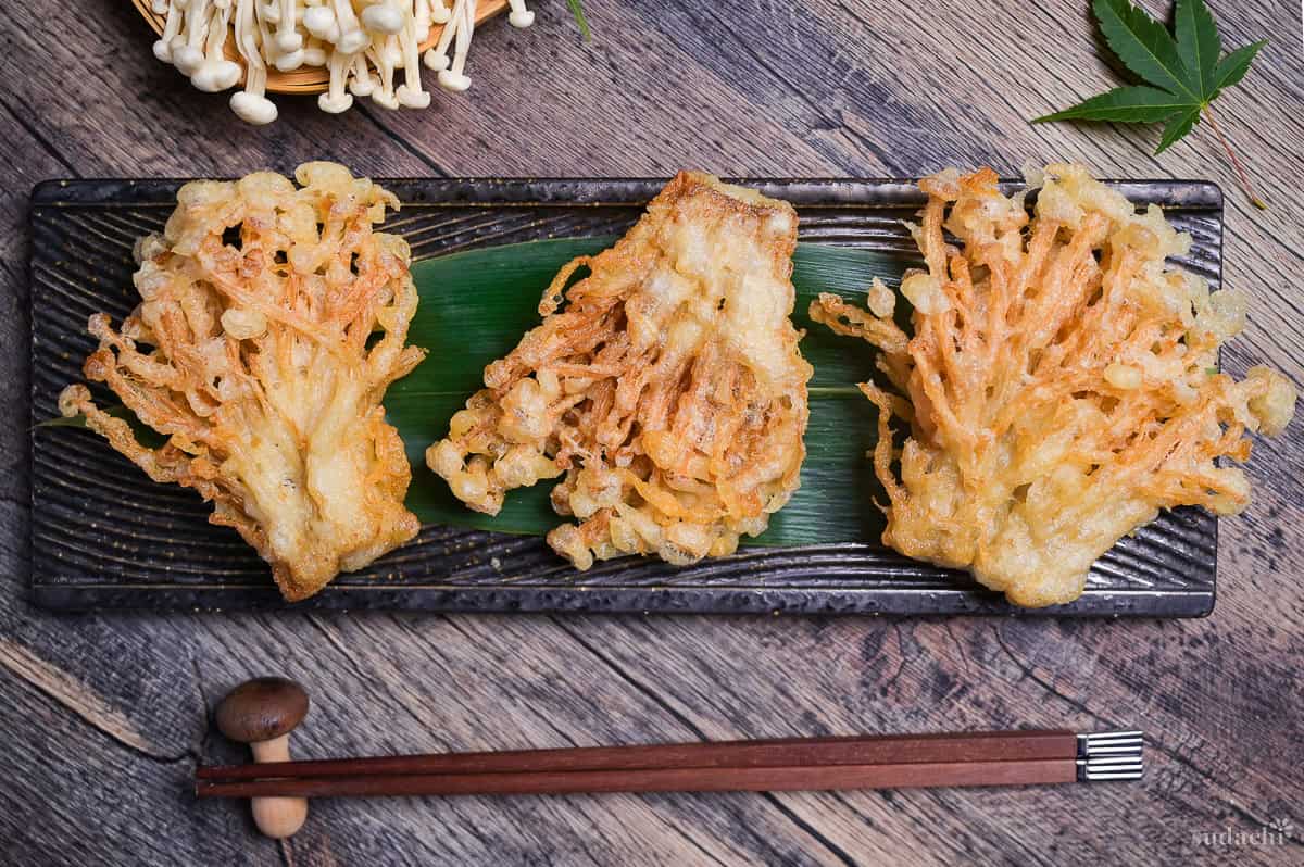 Three bunches of deep fried enoki mushroom tempura arranged over a bamboo leaf on a long black rectangular plate with wavey lines grooves design top down view