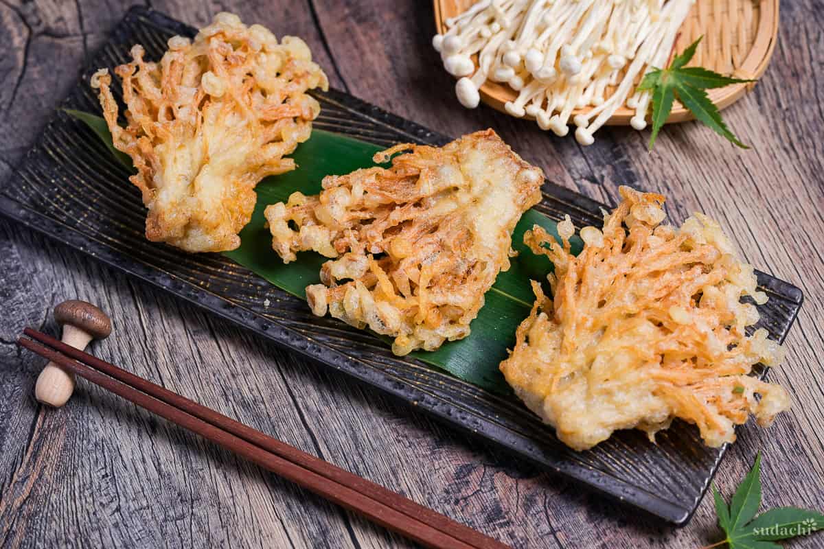 Three bunches of deep fried enoki mushroom tempura arranged over a bamboo leaf on a long black rectangular plate with wavey lines grooves design