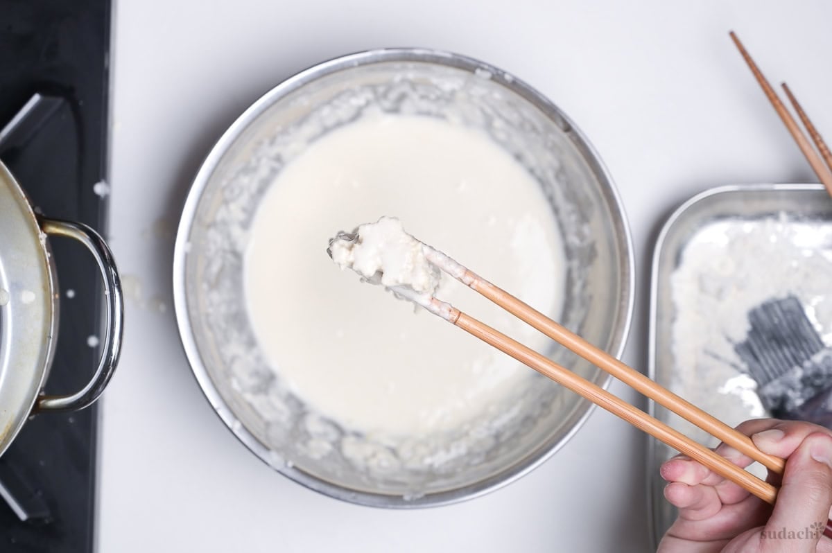 a small bunch of enoki mushrooms coated with tempura batter, held up with wooden cooking chopsticks to let excess drip off