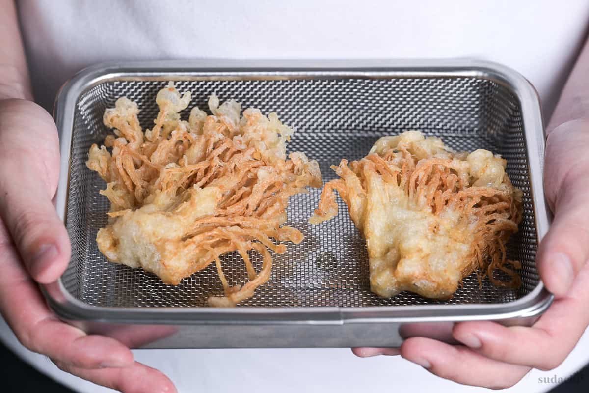Yuto Omura holding up two bunches of enoki mushroom tempura on a mesh wire rack inside a steel container