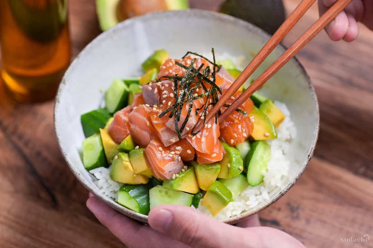 marinated salmon sashimi bowl with cucumber and avocado topped with sesame seeds and kizami nori in a mottled gray ceramic bowl with wooden chopsticks lifting a piece of salmon