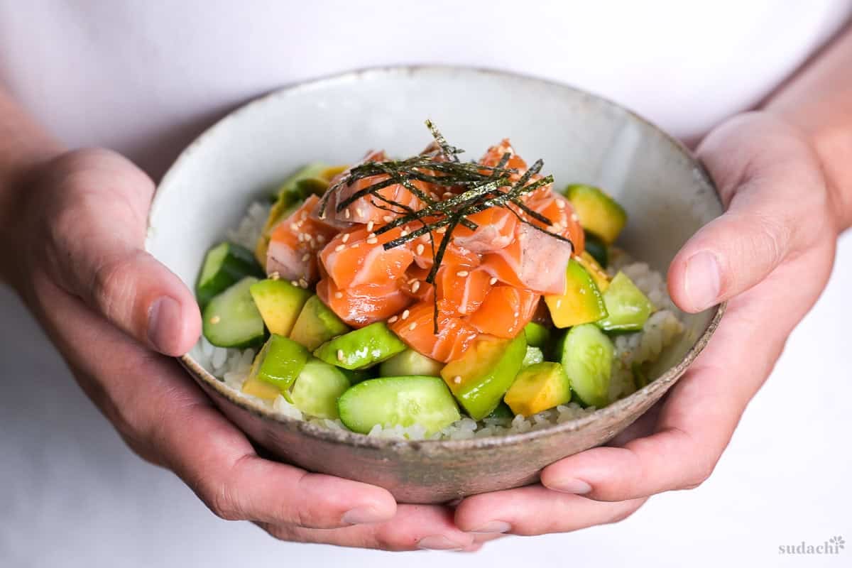 Yuto Omura holding up marinated salmon sashimi bowl with cucumber and avocado topped with sesame seeds and kizami nori in a mottled gray ceramic bowl