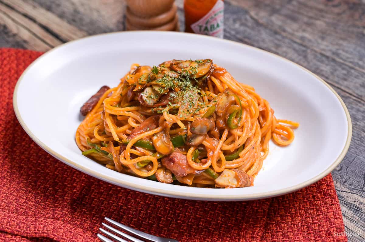 Japanese Ketchup Spaghetti (Spaghetti Napolitan) on a white oval plate on a red woven placemat with pepper grinder and Tabasco chili sauce in the background