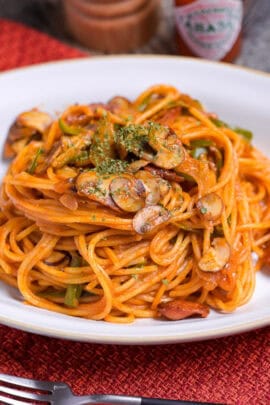 Japanese Ketchup Spaghetti (Spaghetti Napolitan) on a white oval plate on a red woven placemat with pepper grinder and Tabasco chili sauce in the background