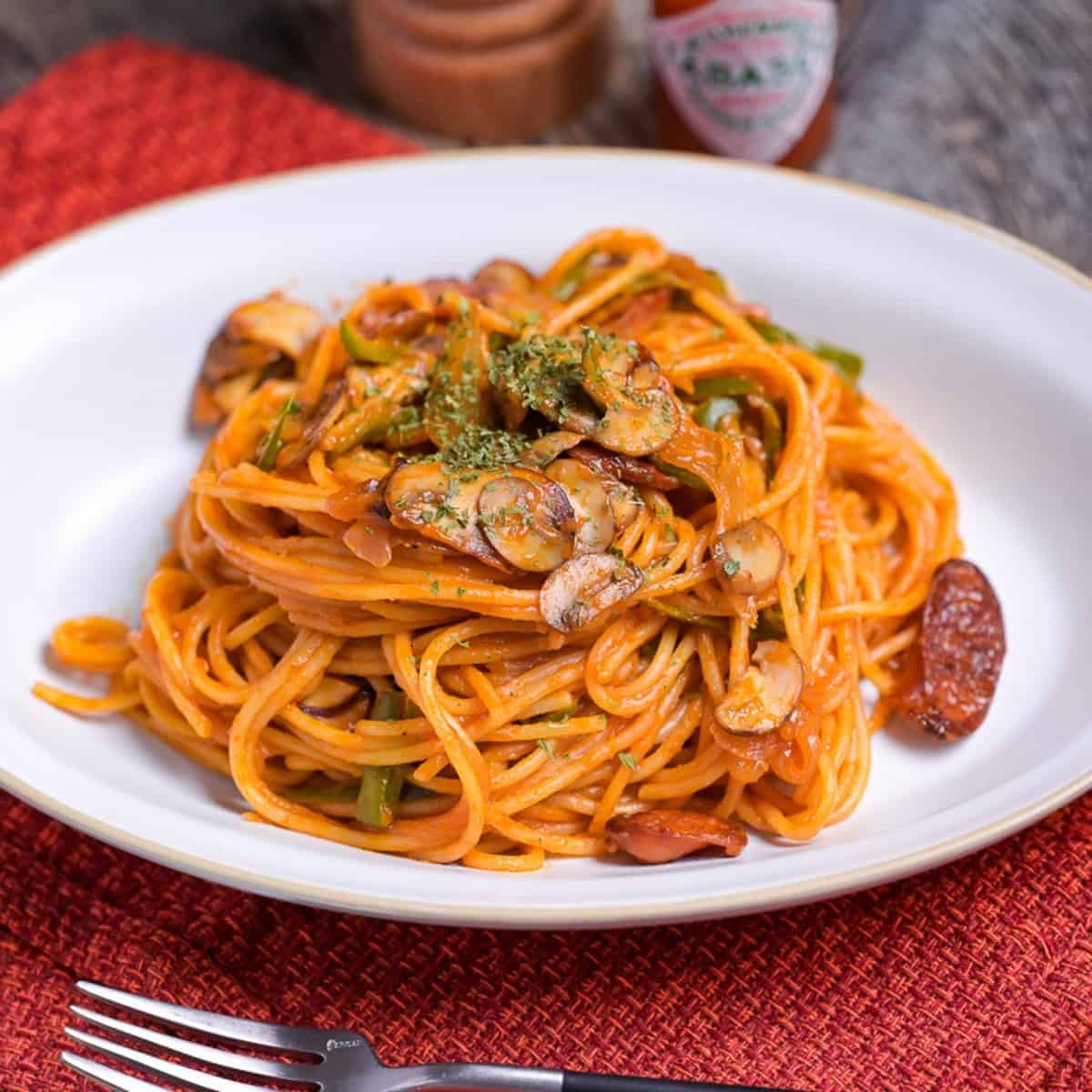 Japanese Ketchup Spaghetti (Spaghetti Napolitan) on a white oval plate on a red woven placemat with pepper grinder and Tabasco chili sauce in the background
