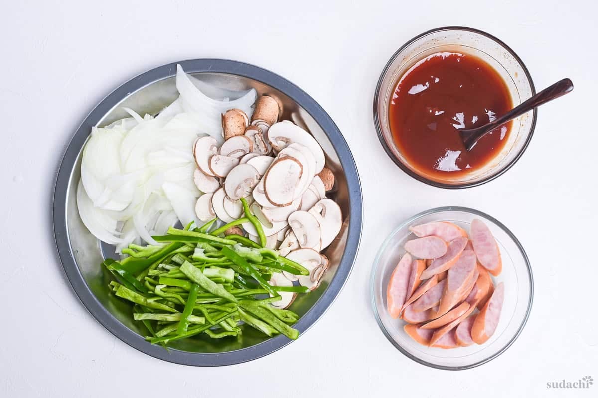 Ingredients prepared for spaghetti napolitan. Sliced onion, green bell pepper and button mushrooms on a plate, sauce mixed in a bowl, and sliced hot dog sausages in another bowl.