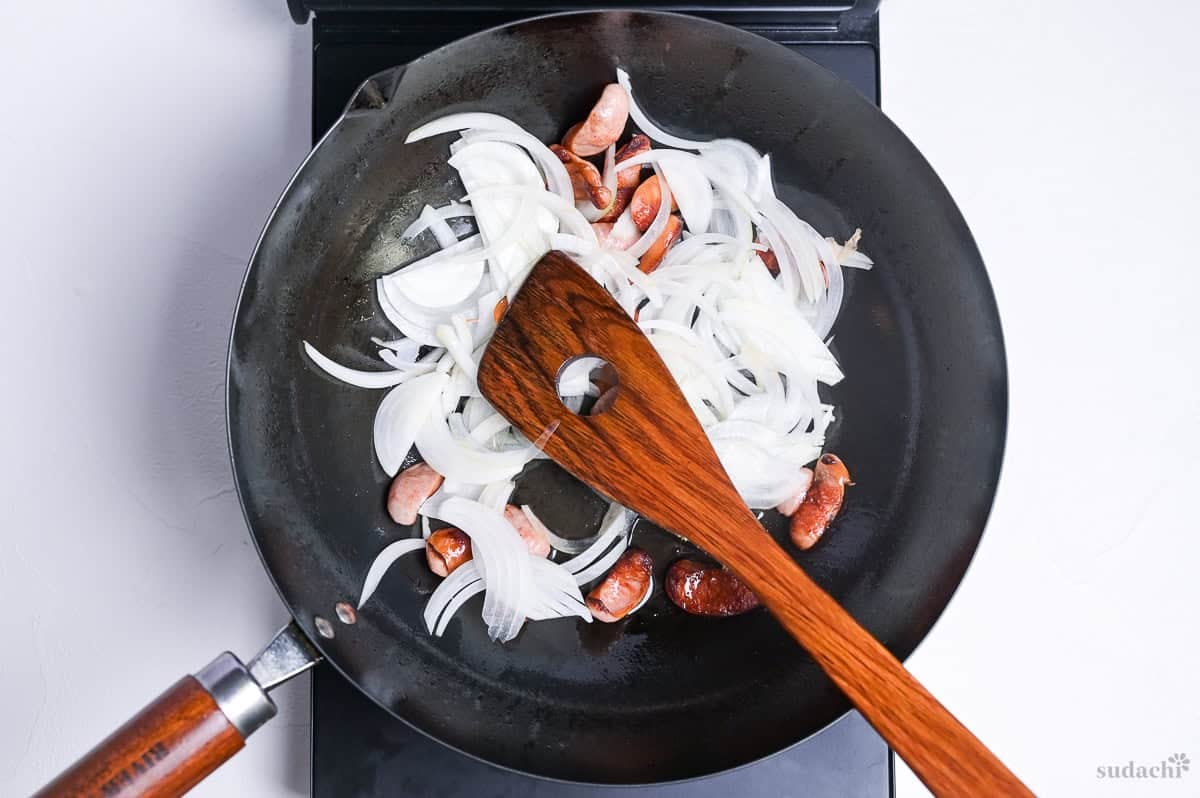 sliced onion and hot dog sausages frying in a wok on the stove top with wooden spatula