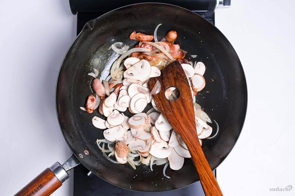 softened onion and seared sausage slices with sliced button mushrooms frying in a wok on the stove top