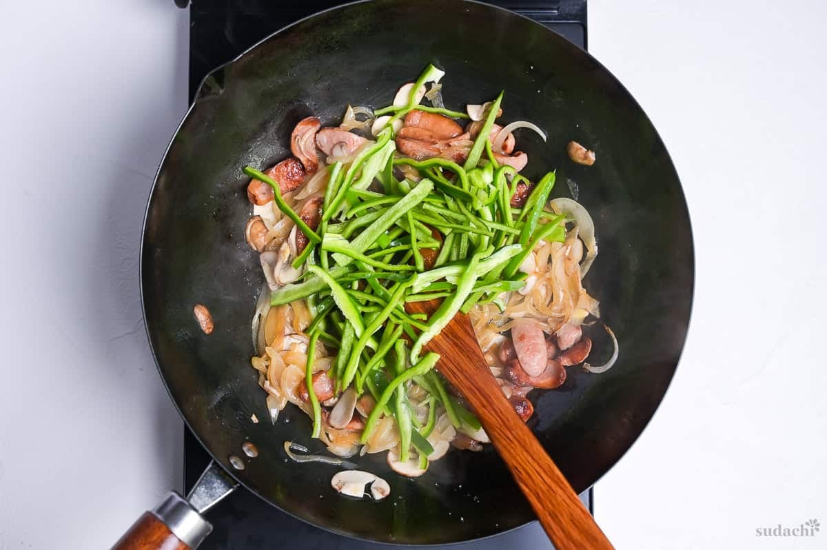 frying hot dog sausages, onion, button mushrooms and green bell pepper in a wok on the stove top.