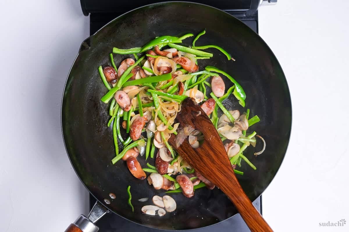 Ingredients for Japanese ketchup spaghetti frying in a wok on the stove top (hot dog sausages, onion, button mushrooms, green bell pepper)