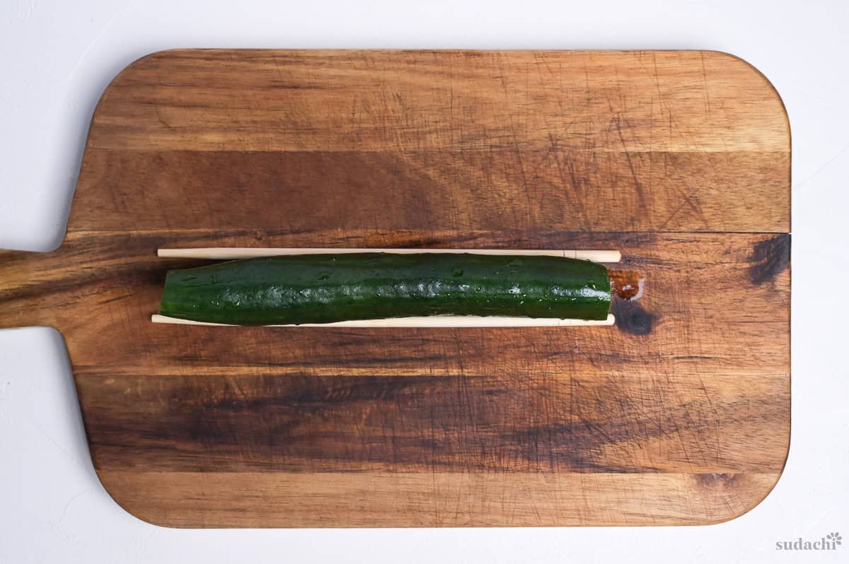 One Japanese cucumber placed between two disposable wooden chopsticks on a wooden cutting board
