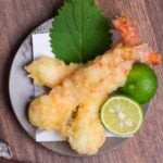Three homemade ebiten (shrimp tempura) arranged on a gray plate next to a halved sudachi and shiso leaf top down