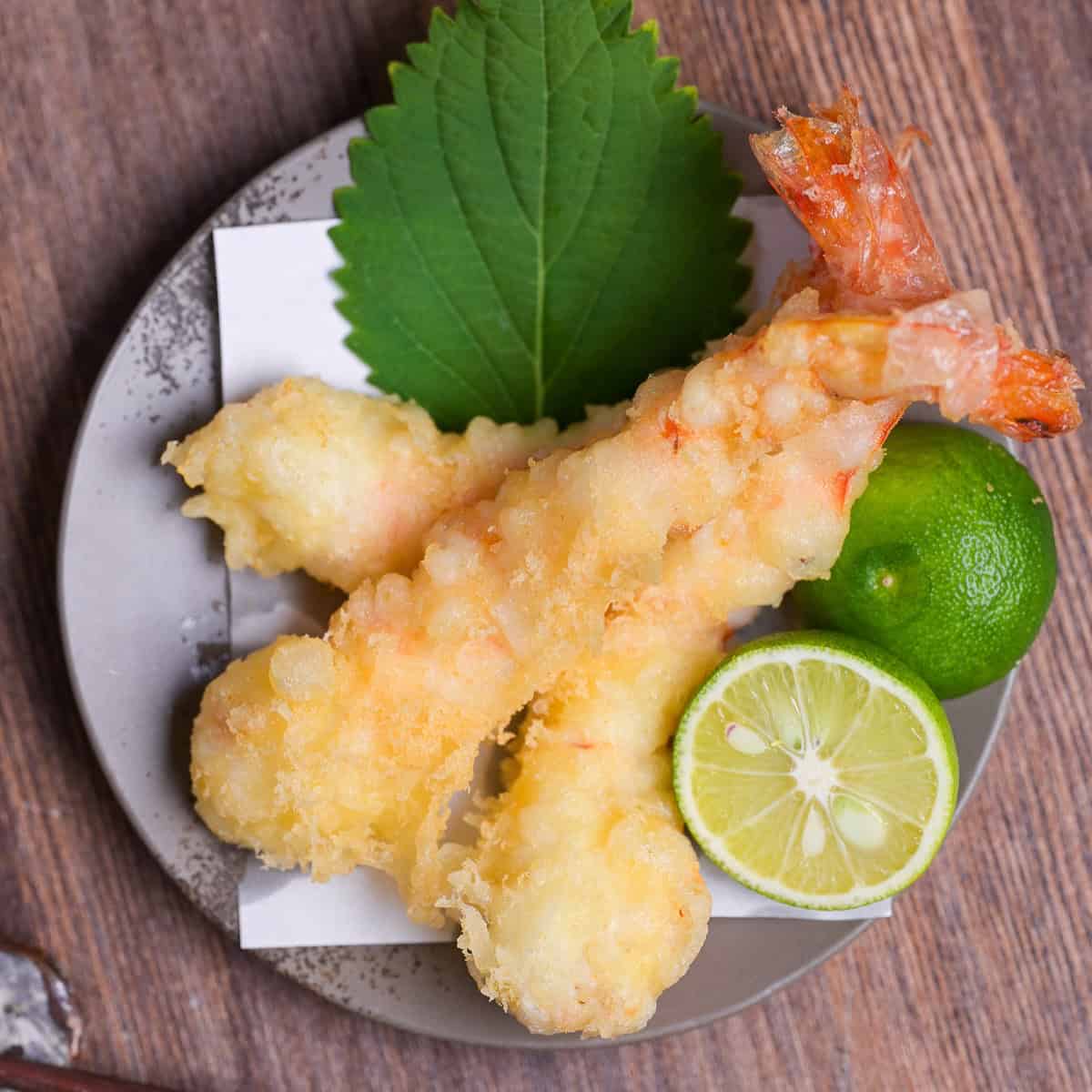 Three homemade ebiten (shrimp tempura) arranged on a gray plate next to a halved sudachi and shiso leaf top down
