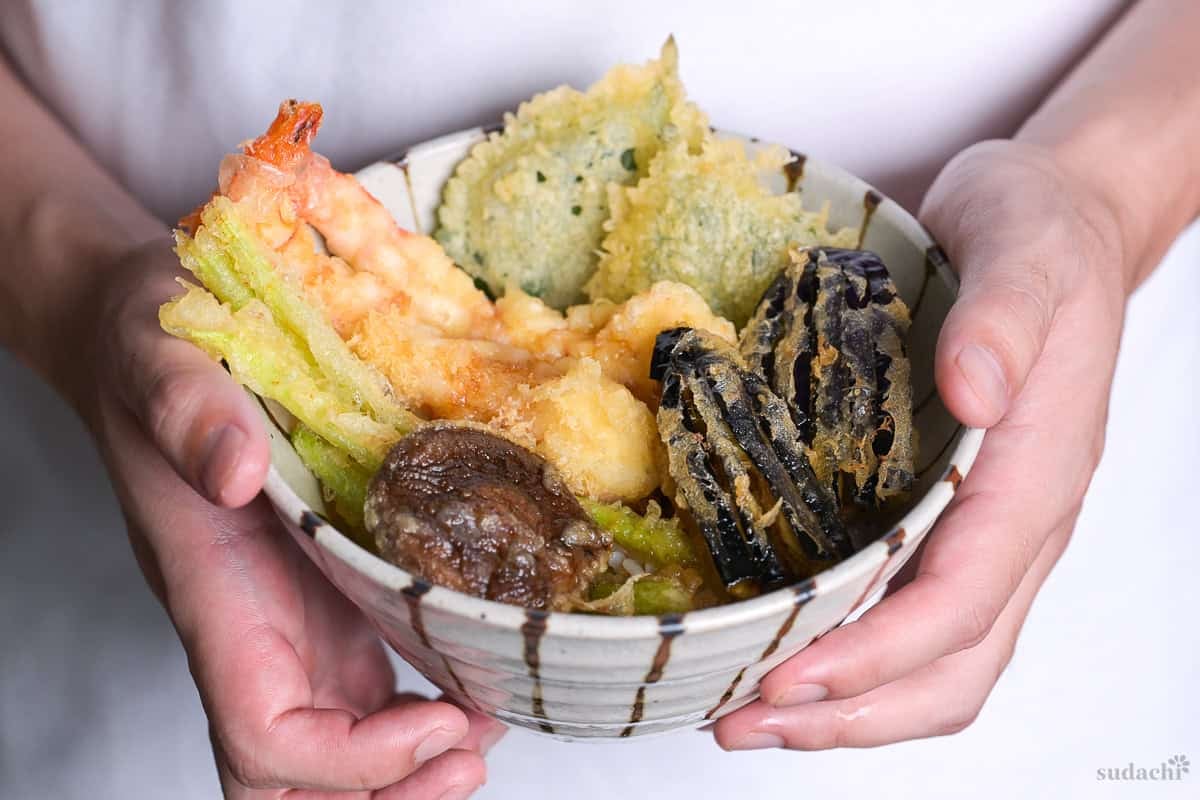 Yuto Omura holding up homemade tempura donburi (tendon) made with shrimp, asparagus, shiso, eggplant and shiitake in a striped bowl.